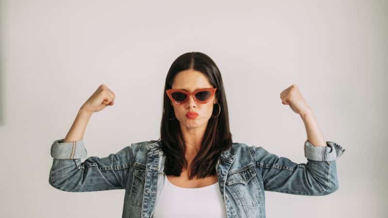 Woman with black hair flexing her arms confidently and looking at the camera. Durchsetzungsvermögen fühlt sich stark an und kann Spaß machen!